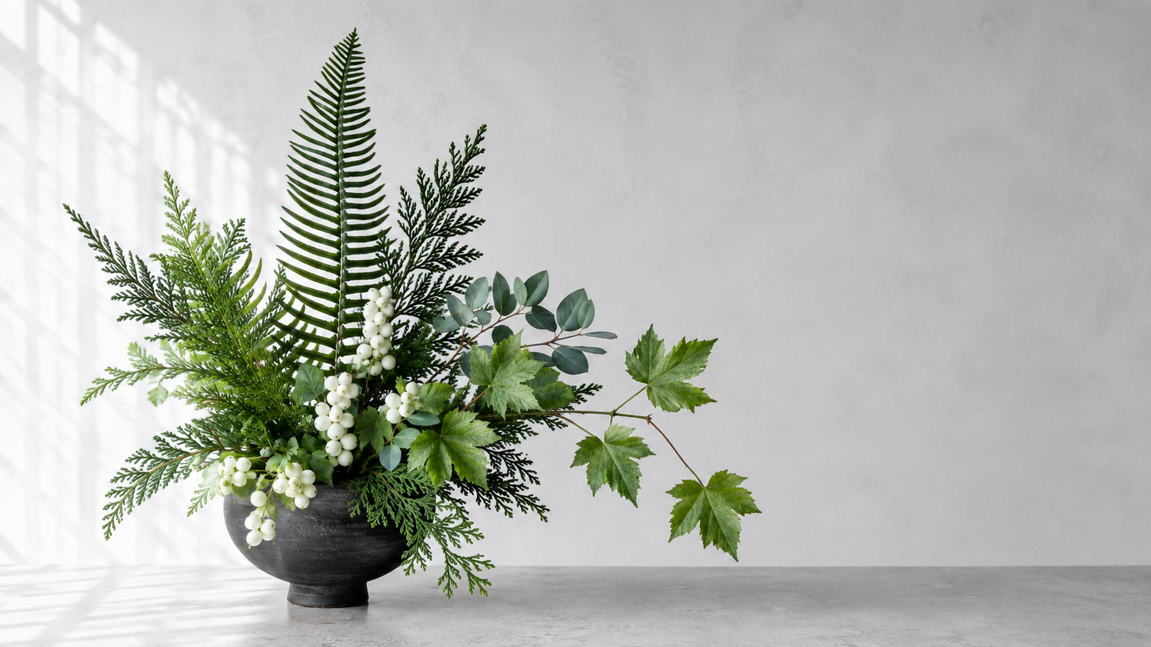 Pacific Northwest woodland foliage for designers. A sculptural woodland arrangement in a dark pedestal vase featuring Western sword fern fronds, cedar tips, eucalyptus, grapevine foliage, and clusters of white snowberry berries.